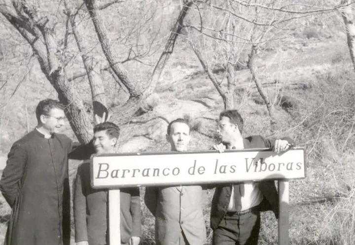 Cuatro hombres, uno con sotana, posan tras un cartel que dice Barranco de las Víboras, entre árboles sin hojas.