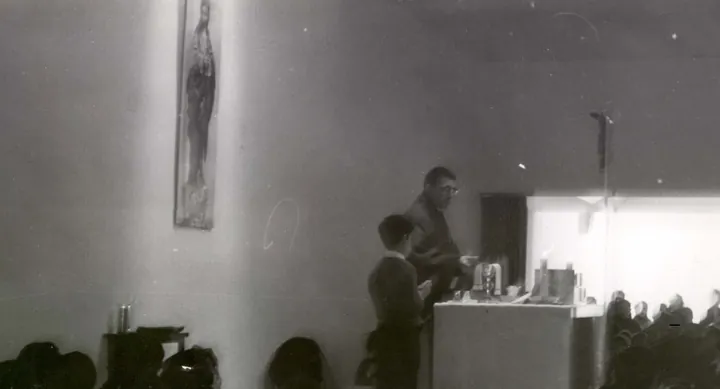 Un hombre ante un altar con cáliz y velas, junto a un niño; en primer plano, cabezas de asistentes.