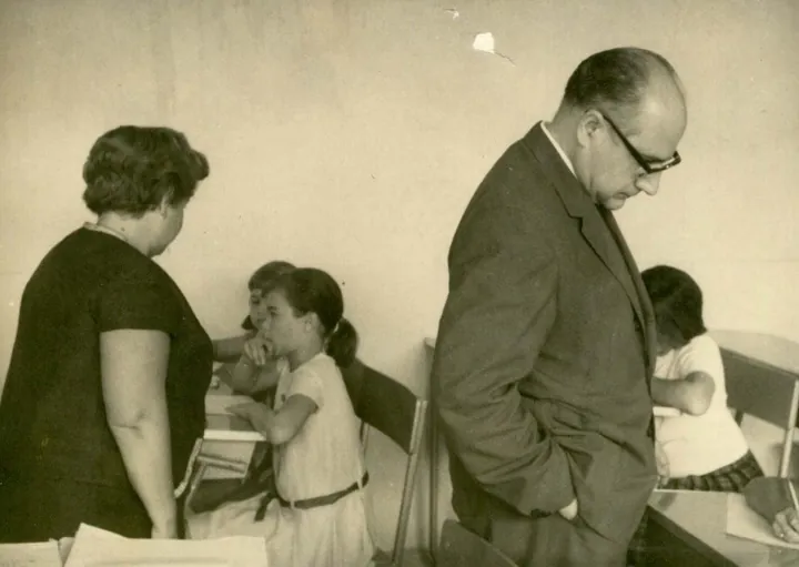 Una mujer de espaldas y un hombre con gafas, de pie, observan a unas niñas sentadas en pupitres.