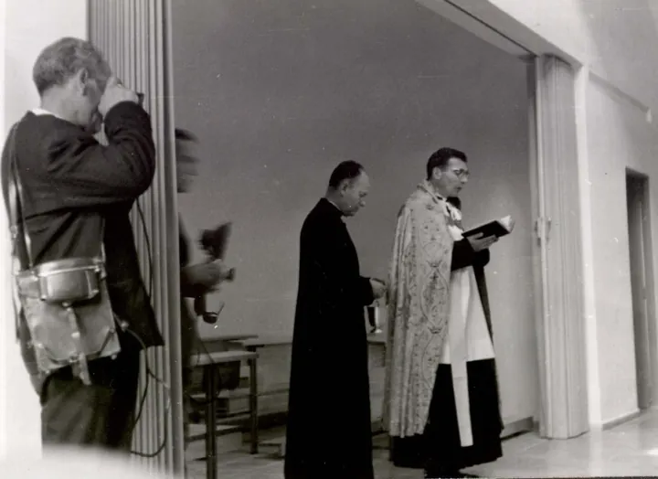 Un sacerdote con casulla lee un libro junto a otro; a la izquierda, un hombre sostiene una cámara.