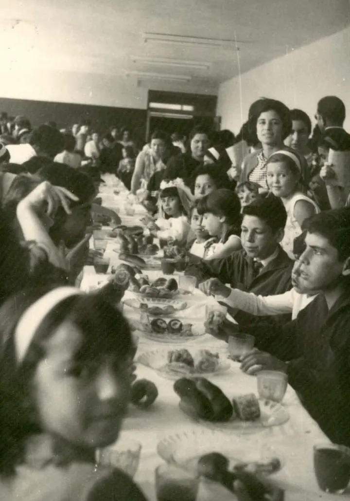 Niños y niñas desayunan sentados a una mesa larga con bollos y vasos, en un comedor lleno.