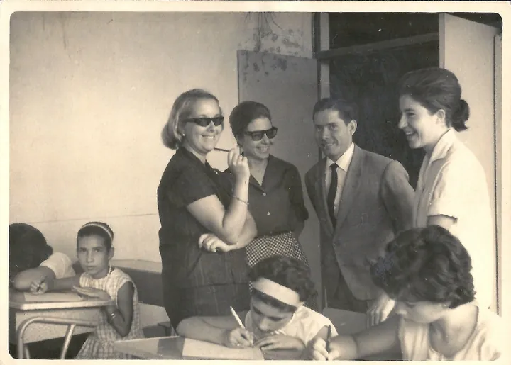 Cuatro adultos sonrientes de pie en un aula, junto a alumnas sentadas que escriben en sus pupitres.