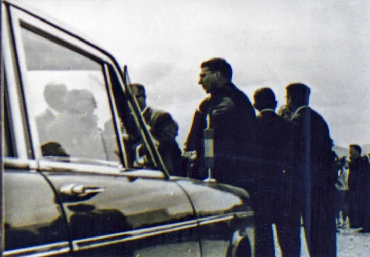 Varios hombres de pie junto a un coche oscuro, a contraluz; en la ventanilla se reflejan otras figuras.