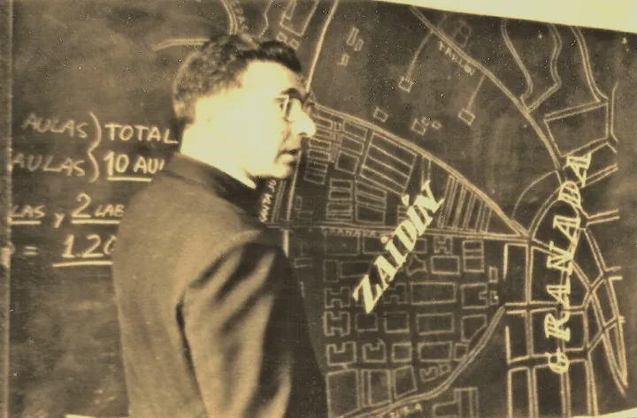 Hombre con gafas, de perfil, ante una pizarra con un plano a tiza donde figuran Zaidín y Granada.