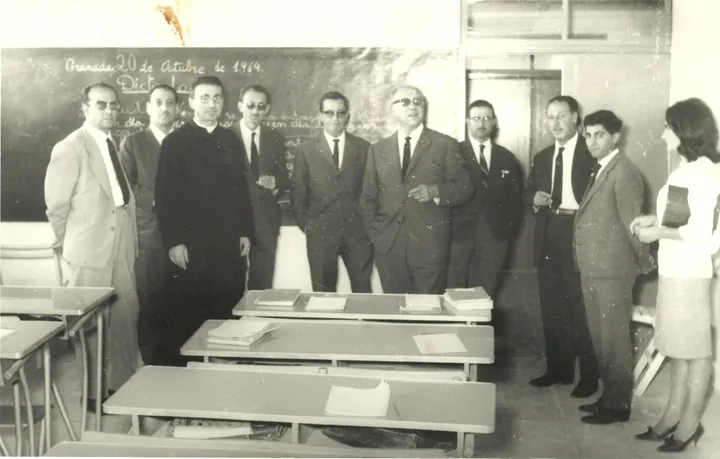Grupo de pie en un aula con pupitres, un sacerdote entre ellos; la pizarra dice 20 de octubre de 1964.