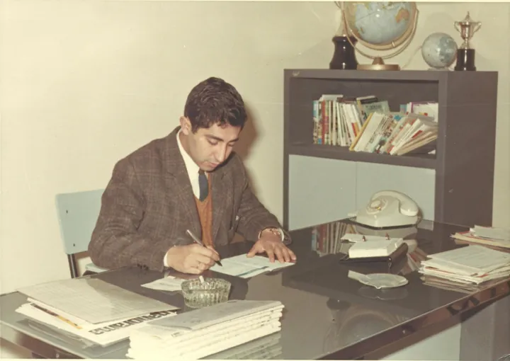 Un hombre con americana escribe en un escritorio; detrás, una estantería con libros, globos terráqueos y un trofeo.